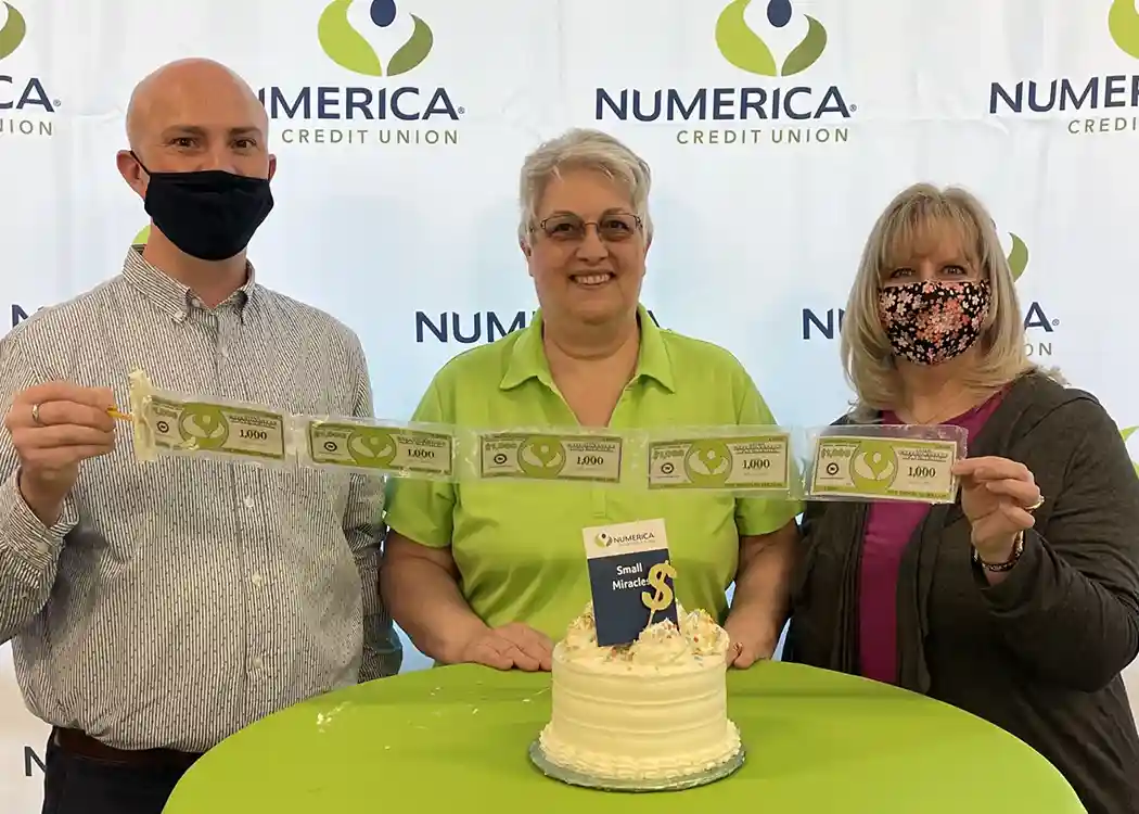 Three people standing in front of a small table with a cake on it, holding a string of play money